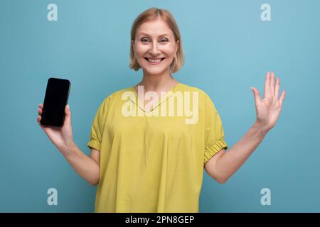 donna bionda di mezza età piacevole che tiene un telefono gadget in mano verticalmente Foto Stock