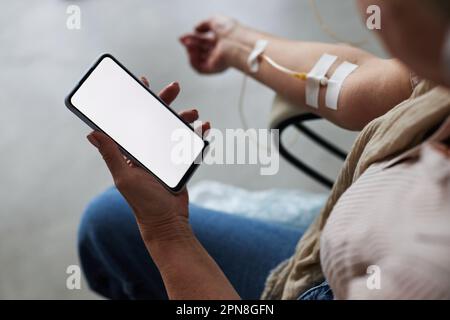 Primo piano di una donna anziana che tiene uno smartphone con schermo vuoto durante il trattamento fleboclisi IV, spazio copia Foto Stock