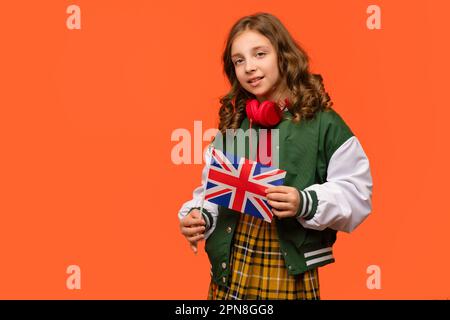 Ragazza adolescente in vestito scuola e cuffie rosse tiene piccola bandiera del Regno Unito. Studio all'estero concetto. Programma di scambio internazionale di studenti. Imparare l'inglese Foto Stock