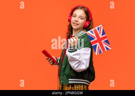 La bambina della scuola tiene una piccola bandiera britannica. Teen in abiti casual e auricolari rossi con telefono cellulare. Corsi di lingue online. Imparare l'inglese britannico. Liste Foto Stock