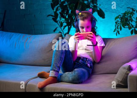 Ragazza adolescente concentrata in cuffie wireless ascoltare musica, utilizzando l'app mobile, navigare sui social media su smartphone in una colorata sala al neon Foto Stock