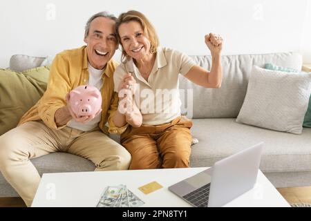 Felice coppia anziana che tiene la banca del piggy e pugni di scuotimento, il calcolo del risparmio urlando fiero Foto Stock