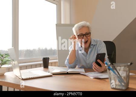 Sicura donna anziana di mezza età euforica vincitrice con smartphone. Signora più anziana matura guardando il telefono delle cellule che legge le notizie grandi ottenendo il buon risultato che vince la sensibilità in linea dell'offerta stupita. Gesto vincente Foto Stock
