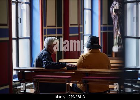 Vista posteriore del sacerdote anziano che parla con il visitatore mentre si siedono sulla panchina durante il ministero in chiesa Foto Stock