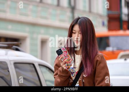Ragazza asiatica con capelli viola tinti camminare con smartphone su una strada. Uso del telefono cellulare nella città primaverile Foto Stock