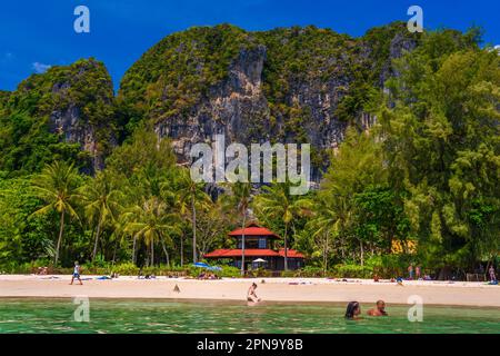 Bungalow casa con tetto rosso tra palme da cocco vicino alle scogliere con persone che prendono il sole e nuotare in acqua smeraldo sulla spiaggia Railay ovest, Ao Nang, K Foto Stock