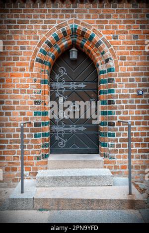 Retro porta in legno con gnocciola in ferro ornato, maniglia e cerniere in ingresso ad arco con mattoni colorati possibile ingresso laterale di una piccola chiesa, cappella Foto Stock