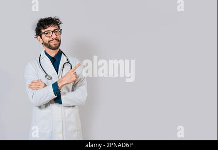 Medico sorridente che punta il dito isolato. Medico che indica uno spazio bianco isolato. Medico giovane che punta il dito in uno spazio vuoto Foto Stock