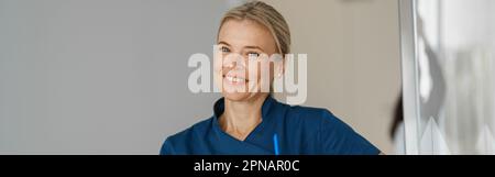 Medico sorridente con appunti in piedi vicino alla reception nella sala clinica e guardando la macchina fotografica Foto Stock