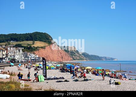 Turisti che si rilassano sulla spiaggia con vista verso gli edifici della città e le scogliere a Pennington Point, Sidmouth, Devon, Regno Unito. Foto Stock