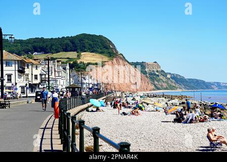 Turisti che si rilassano sulla spiaggia con vista verso gli edifici della città e le scogliere a Pennington Point, Sidmouth, Devon, Regno Unito. Foto Stock