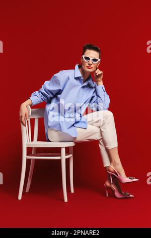 Donna adulta in occhiali da sole, camicia e pantaloni siede in profilo sulla sedia e guarda direttamente in avanti Foto Stock