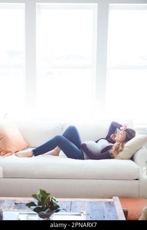 Prendere un pisolino con il bambino. Scatto a tutta lunghezza di una giovane donna incinta attraente che prende un pisolino sul divano a casa. Foto Stock