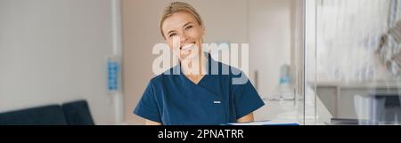 Medico sorridente con appunti in piedi vicino alla reception nella sala clinica e guardando la macchina fotografica Foto Stock