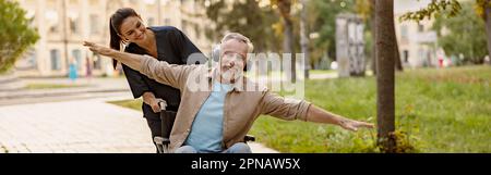 Gioioso uomo disabili maturo in sedia a rotelle indossando le cuffie divertirsi durante una passeggiata in città assistita da una bella giovane infermiera Foto Stock