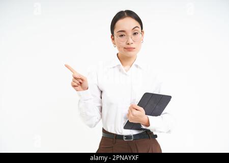 Ritratto di donna corporate coreana in occhiali, tiene il tablet digitale, punta il dito a sinistra e appare severo e serio, si trova su sfondo bianco Foto Stock
