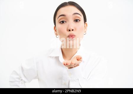 Primo piano ritratto di carina giovane donna asiatica, soffia aria bacio con labbra puckered, manda mwah alla macchina fotografica, in piedi su sfondo bianco Foto Stock