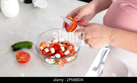 Una ragazza incinta prepara un'insalata di verdure di cetrioli e pomodori. Il concetto di nutrizione delle donne incinte con vitamine e macronutrienti in v Foto Stock