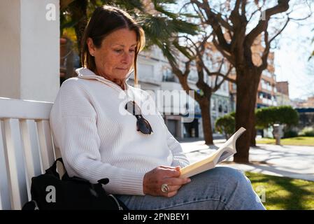 Donna seria di mezza età in abiti casual seduti su panca e libro di lettura mentre riposa nel parco nella giornata di sole Foto Stock