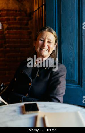 Donna di mezza età positiva in giacca nera guardando la fotocamera mentre si siede al tavolo con smartphone vicino alla porta blu Foto Stock