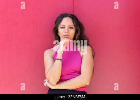 Ritratto di donna penosa con lunghi capelli scuri in alto in piedi vicino alla parete rosa e toccare il mento mentre si guarda la fotocamera Foto Stock