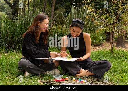 Felice giovane multietnica amici donne seduto su erba e disegno con coloratissime pitture acquerello mentre si passa il tempo in natura Foto Stock