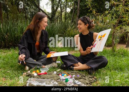 Felice giovane multietnica amici donne seduto su erba e disegno con coloratissime pitture acquerello mentre si passa il tempo in natura Foto Stock