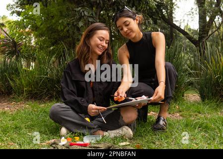 Felice giovane multietnica amici donne seduto su erba e disegno con coloratissime pitture acquerello mentre si passa il tempo in natura Foto Stock