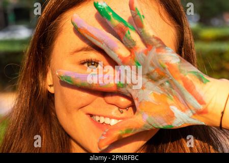 Primo piano di allegra giovane artista femminile che chiude l'occhio con la mano cosparsa di colori luminosi mentre si guarda la fotocamera su sfondo sfocato Foto Stock