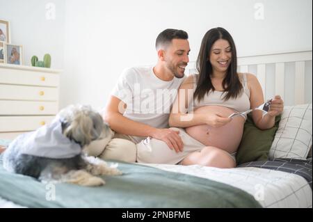 Giovane uomo allegro con donna incinta sorridente e guardando la foto ecografica del bambino mentre seduto sul letto vicino al cane in camera da letto accogliente Foto Stock