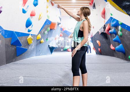Vista laterale di una giovane donna in piedi vicino alla parete di bouldering con impugnature codificate per colore e dito di puntamento mentre si guarda in alto nella palestra di allenamento per arrampicata su roccia Foto Stock