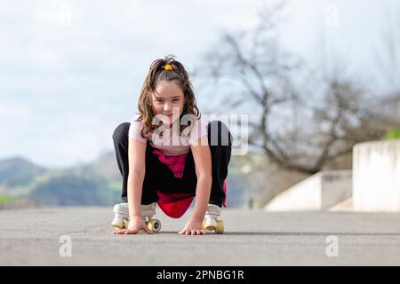 Corpo pieno di bambina seduta con le mani su strada asfaltata e guardando la macchina fotografica mentre pattina con pattini a rotelle in parco sfocato durante il giorno Foto Stock