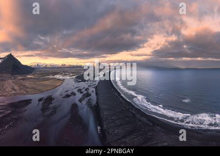 Vista aerea delle montagne rocciose coperte di neve situato vicino al mare contro il cielo nuvoloso durante il tramonto in Islanda Foto Stock