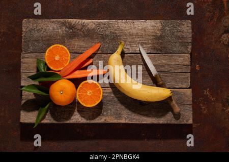 Vista dall'alto disposizione di arance mature e banana poste vicino a fette di carota tagliata con coltello su superficie di legno Foto Stock