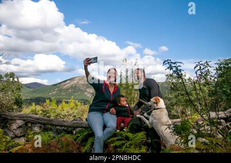 Felice giovane famiglia mista corsa seduta sul tronco di albero di legno secco e prendendo selfie sul telefono cellulare, mentre guardando lo schermo e rilassarsi nella natura con Foto Stock