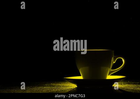 Una tazza con una bevanda calda illuminata da luce gialla su sfondo nero, silhouette, spazio copia, creativo. Caffè mattutino Foto Stock