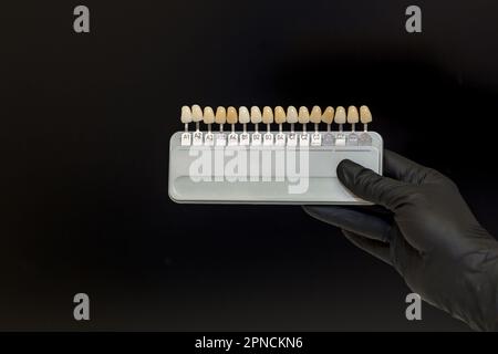 La mano di un dentista in un guanto nero contiene una tavolozza di colori con denti artificiali per una selezione visiva dei colori su sfondo nero Foto Stock