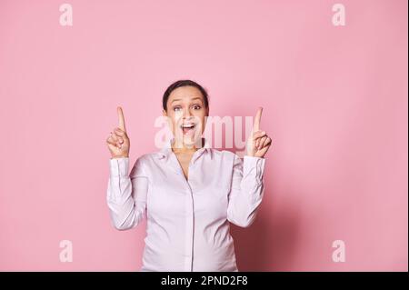 Donna incinta stupita che punta le dita verso l'alto, in posa con la bocca aperta, esprimendo emozioni positive su sfondo rosa Foto Stock