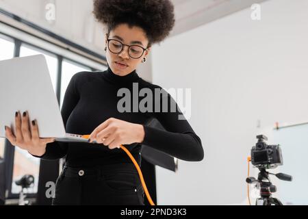african american content manager in occhiali e dolcevita nero cavo di collegamento al computer portatile in studio fotografico, immagine stock Foto Stock
