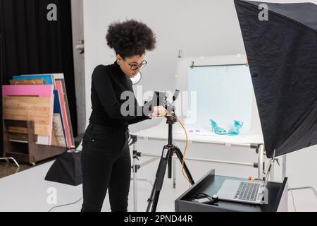 produttore di contenuti afroamericano che collega la fotocamera digitale al computer portatile in un moderno studio fotografico, immagine di scorta Foto Stock