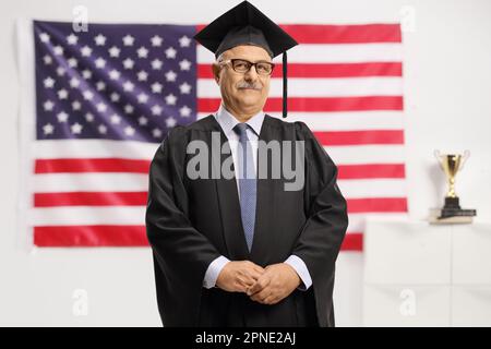 Uomo maturo che indossa un abito da laurea e che si erge di fronte a una bandiera degli Stati Uniti isolata su sfondo bianco Foto Stock