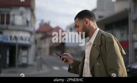 Un giovane del Medio Oriente che cammina per strada in città guardando il dispositivo cellulare. Un maschio arabo che controlla i contenuti online in ambiente urbano Foto Stock