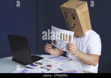 Un uomo è un uomo d'affari in una camicia bianca con un sacchetto di carta sulla sua testa, con un volto sorridente gioioso disegnato, conduce una video conferenza o l'addestramento via vid Foto Stock