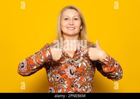 Donna bionda caucasica nel suo 40s in un abito modellato che indica a se stessa orgogliosa di essere il modo in cui è, donna forte e indipendente isolato sopra Foto Stock