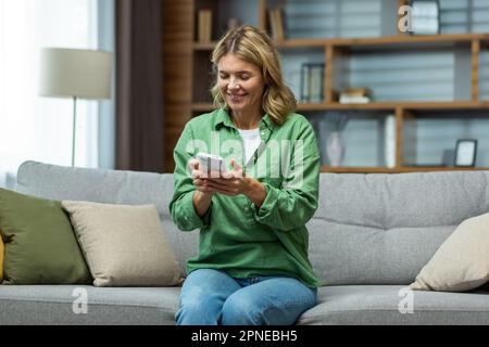 Donna anziana felice seduta sul divano a casa e utilizzando il telefono. Parla, legge messaggi, gioca a giochi online. Foto Stock