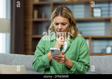 Donna anziana sconvolta seduta sul divano a casa e tenendo il telefono. Guarda lo schermo in modo preoccupante, ha ricevuto un messaggio, cattive notizie. Foto Stock