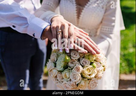 Anelli di nozze sulle mani degli sposi novelli, un bouquet di fiori sullo sfondo. Anelli d'oro sulle mani di un uomo e di una donna Foto Stock