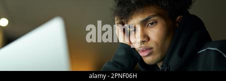 Ritratto di studente maschile annoiato che studia, lavora su un computer portatile, seduto in camera oscura a casa a tarda notte Foto Stock