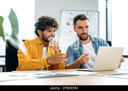 Due dipendenti professionisti multirazziali di sesso maschile siedono in ufficio al tavolo, guardano lo schermo del notebook, lavorano a un progetto comune, si consultano, discutono idee e strategie, valutano i rischi Foto Stock
