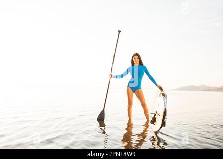 Donna che indossa costume da bagno blu, con un remo e una tavola da paddleboard in lago Foto Stock